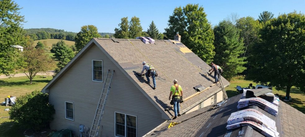 Roofers repairing a shingle roof on a suburban house with ladders and roofing materials nearby
